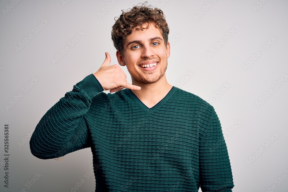 Young blond handsome man with curly hair wearing green sweater over white background smiling doing phone gesture with hand and fingers like talking on the telephone. Communicating concepts.