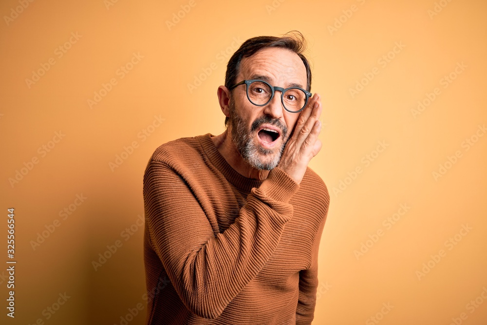 Middle age hoary man wearing brown sweater and glasses over isolated yellow background hand on mouth telling secret rumor, whispering malicious talk conversation Middle age hoary man wearing brown sweater and glasses over isolated yellow background hand on mouth telling secret rumor, whispering malicious talk conversation