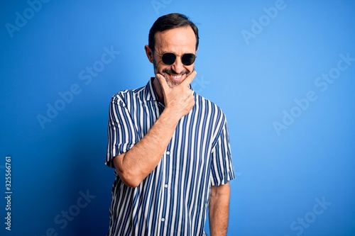 Middle age hoary man wearing striped shirt and sunglasses over isolated blue background looking confident at the camera smiling with crossed arms and hand raised on chin. Thinking positive.