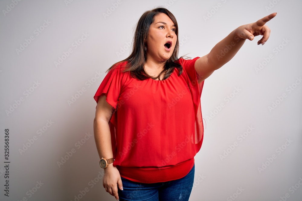Beautiful brunette plus size woman wearing casual red t-shirt over isolated white background Pointing with finger surprised ahead, open mouth amazed expression, something on the front