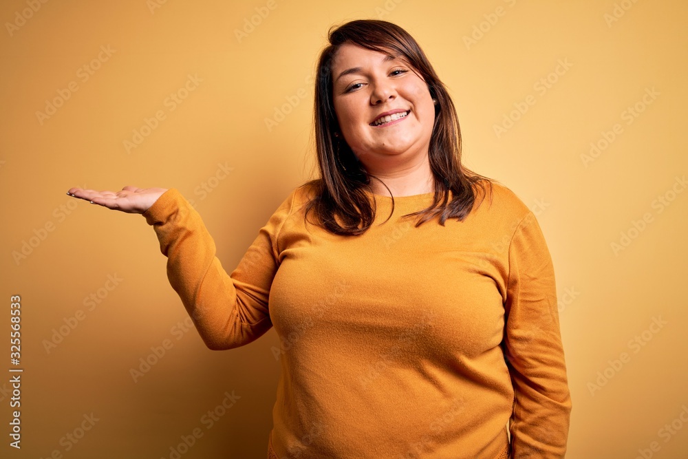 Beautiful brunette plus size woman wearing casual sweater over isolated yellow background smiling cheerful presenting and pointing with palm of hand looking at the camera. Beautiful brunette plus size woman wearing casual sweater over isolated yellow background smiling cheerful presenting and pointing with palm of hand looking at the camera.