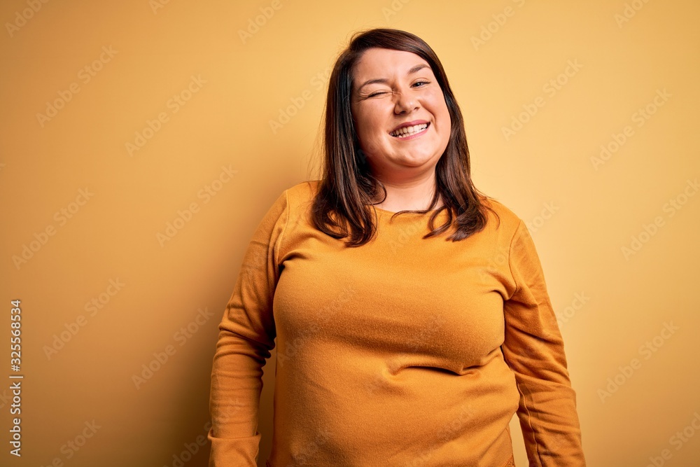 Obraz premium Beautiful brunette plus size woman wearing casual sweater over isolated yellow background winking looking at the camera with sexy expression, cheerful and happy face.