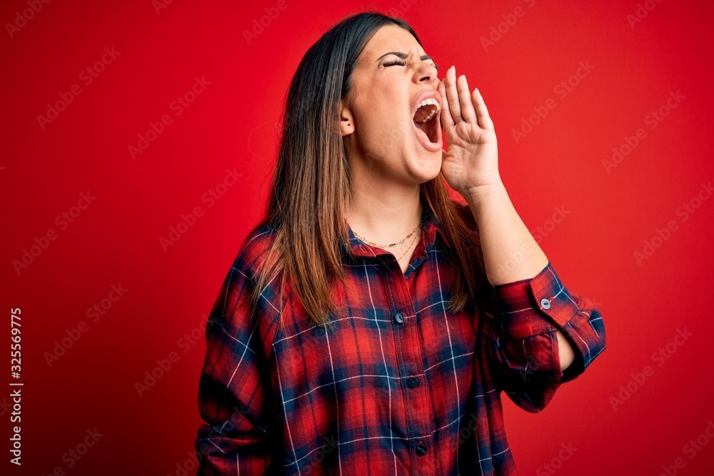 Young beautiful woman wearing casual shirt over red background shouting and screaming loud to side with hand on mouth. Communication concept. Young beautiful woman wearing casual shirt over red background shouting and screaming loud to side with hand on mouth. Communication concept.