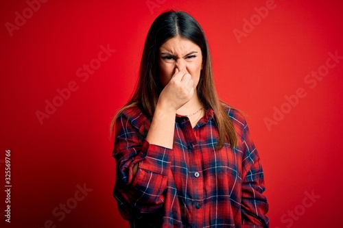 Young beautiful woman wearing casual shirt over red background smelling something stinky and disgusting, intolerable smell, holding breath with fingers on nose. Bad smell © Krakenimages.com