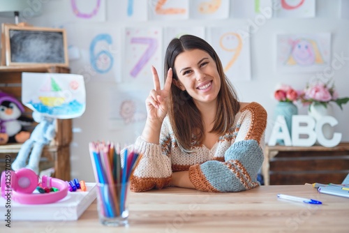 Young beautiful teacher woman wearing sweater and glasses sitting on desk at kindergarten smiling with happy face winking at the camera doing victory sign. Number two.