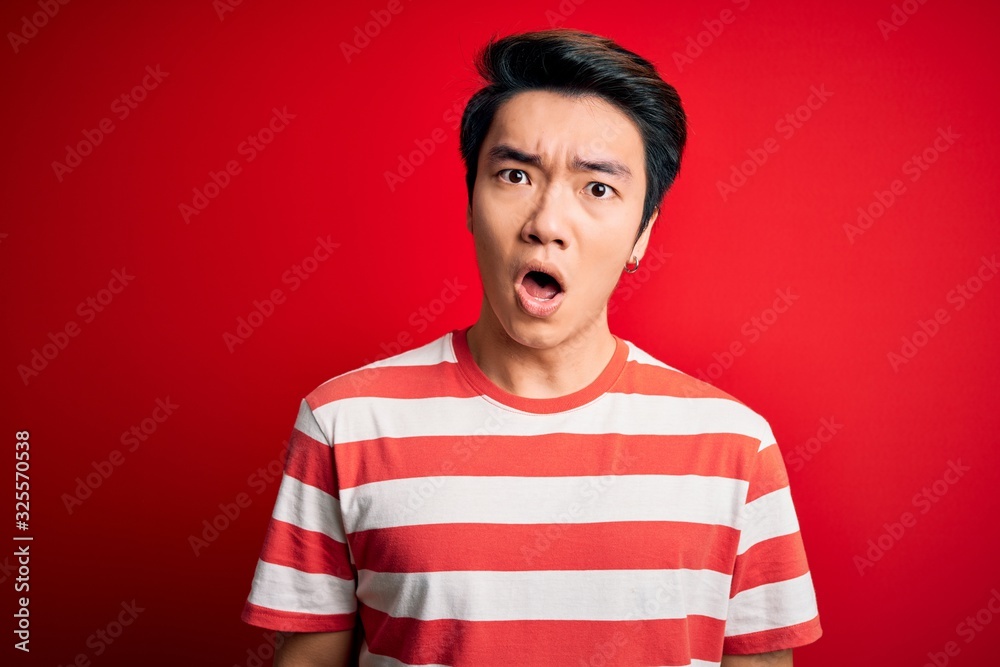 Young handsome chinese man wearing casual striped t-shirt standing over red background In shock face, looking skeptical and sarcastic, surprised with open mouth Young handsome chinese man wearing casual striped t-shirt standing over red background In shock face, looking skeptical and sarcastic, surprised with open mouth