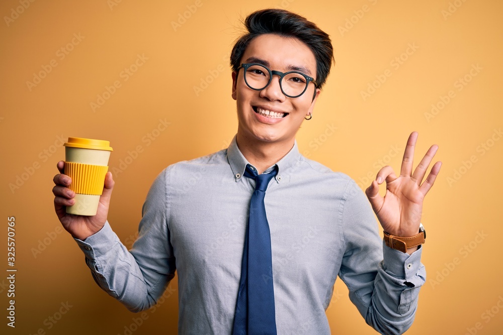 Young handsome chinese man drinking glass of take away coffee over yellow background doing ok sign with fingers, excellent symbol Young handsome chinese man drinking glass of take away coffee over yellow background doing ok sign with fingers, excellent symbol