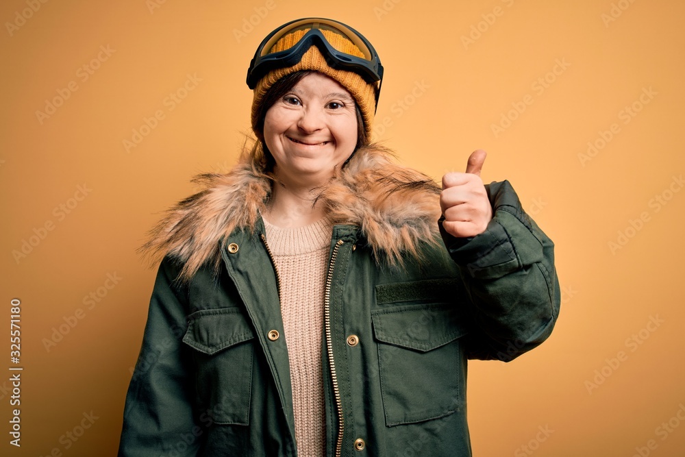Young down syndrome woman wearing ski coat and glasses for winter weather doing happy thumbs up gesture with hand. Approving expression looking at the camera showing success.