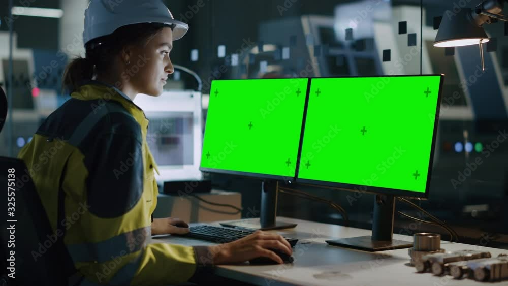 Female Engineer Wearing Safety Vest and Hardhat Works on a Computer, Two Monitor Screens Show Chroma Key / Green Screen Displays. Modern High-Tech Industry 4.0 Electronics Production Factory