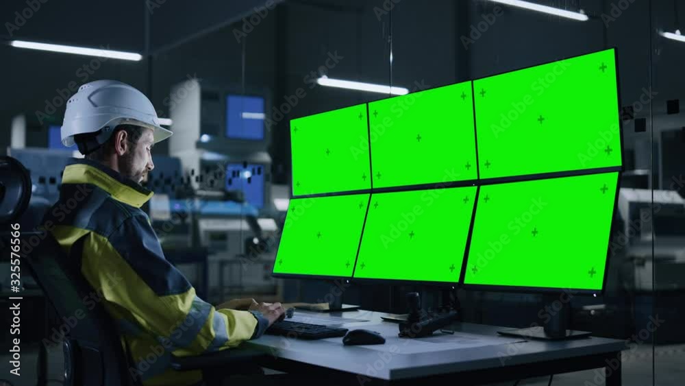 Industry 4.0 Modern Factory: Handsome Male Operator Controls Facility, Uses Computer with Multiple Chroma Key / Green Screen Displays. In Background Dark Automated Electronics Producing Facility