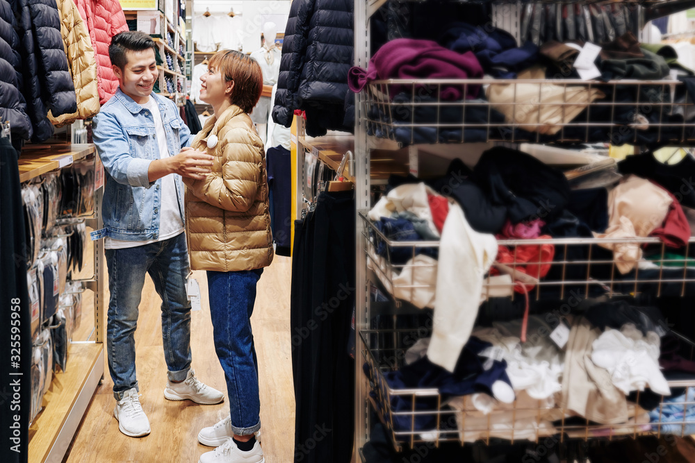 Cheerful young man helping girlfriend to zip up winter jacket she is trying on in store