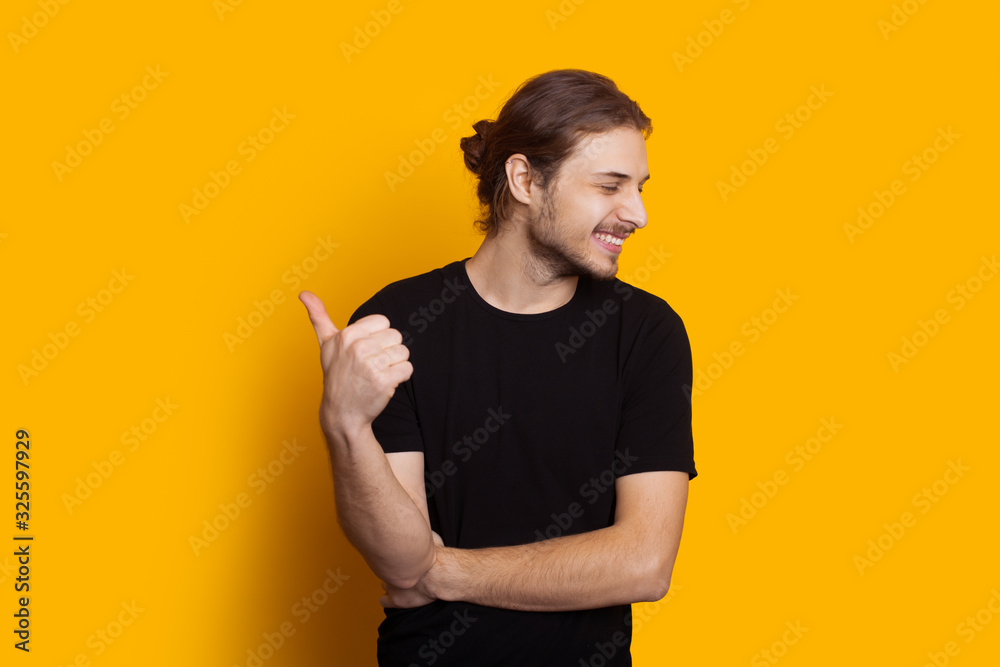 Dressed in black caucasian man with long hair and beard is pointing up while smiling and posing on a yellow background