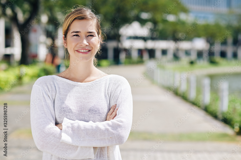 Lovely mixed-race young woman in white sweater standing in city park with arms folded and looking at camera Lovely mixed-race young woman in white sweater standing in city park with arms folded and looking at camera