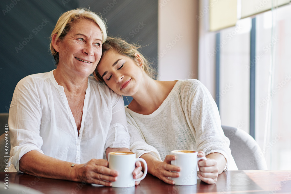 Smiling charming young woman leaning on shoulder of her mother when they are drinking coffee together