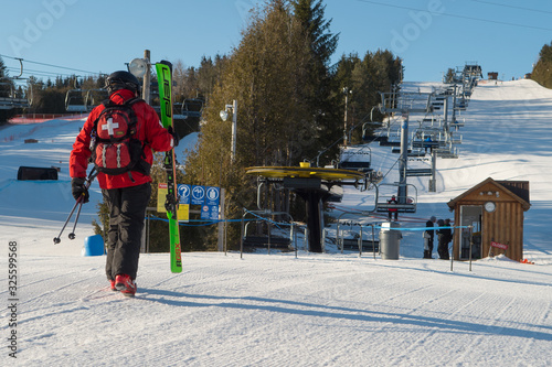 Ski Patrol on Ski Hill walking towards ski chairlift carrying green skis on blue sky day wearing ski patrol uniform with white cross on back horizontal format room for type