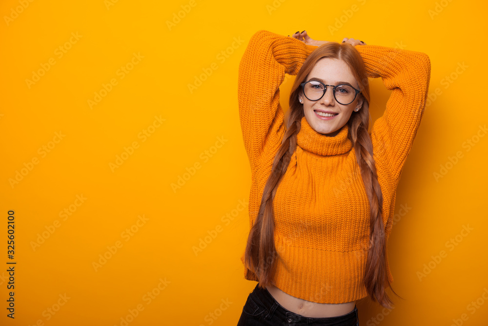 Charming red haired student with red hair and glasses is stretching her hands while posing in an orange sweater on a yellow background Charming red haired student with red hair and glasses is stretching her hands while posing in an orange sweater on a yellow background