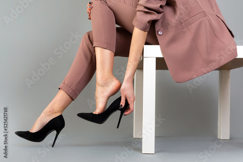 The concept of varicose disease. A woman in a suit takes off her high-heeled shoes. Legs close up. Gray background