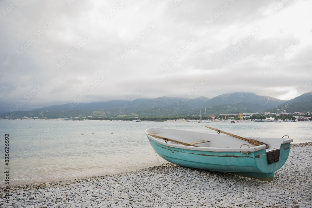 side view of small wooden fishing azure boat on pebble coast black sea beach in bad weather on dramatic sea, sky, mountain and town background, horizontal stock photo image