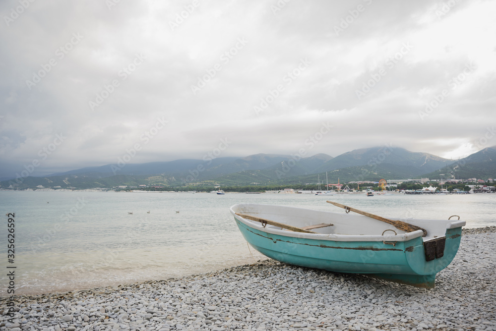 side view of small wooden fishing azure boat on pebble coast black sea beach in bad weather on dramatic sea, sky, mountain and town background, horizontal stock photo image side view of small wooden fishing azure boat on pebble coast black sea beach in bad weather on dramatic sea, sky, mountain and town background, horizontal stock photo image