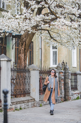 young stylish woman walking by city street in coat blooming tree on background