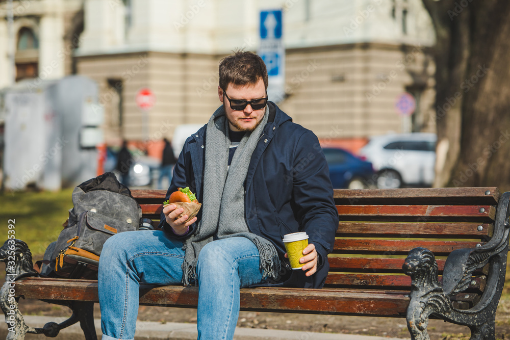 young smiling man eating burger and drink coffee sitting on city bench young smiling man eating burger and drink coffee sitting on city bench