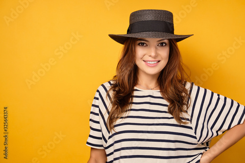 Charming brunette in a striped T-shirt stands on a yellow background in a hat and smiles.
