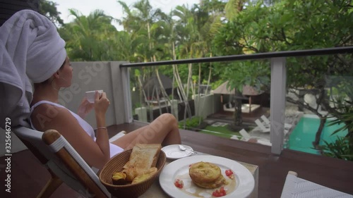 nice woman in bikini and towel on her head rests on hotel belcony with cup of coffee in hand and pastry on table enjoying warm morning photo
