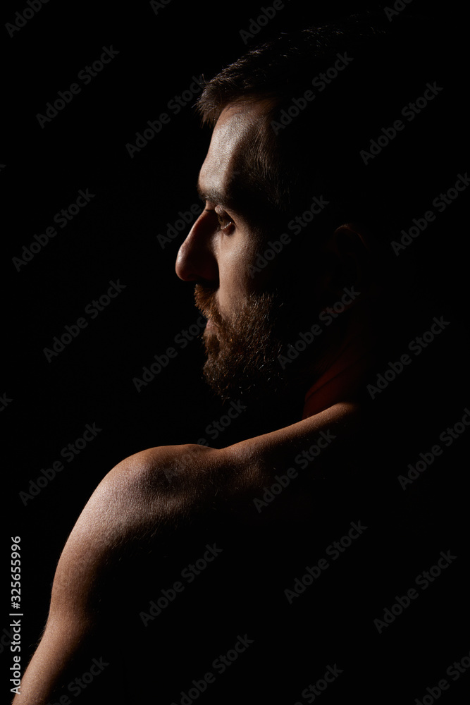 Caucasian young man with beard, no shirt, muscular body, looking away, silhouette, artistic pose, on black background, vertical