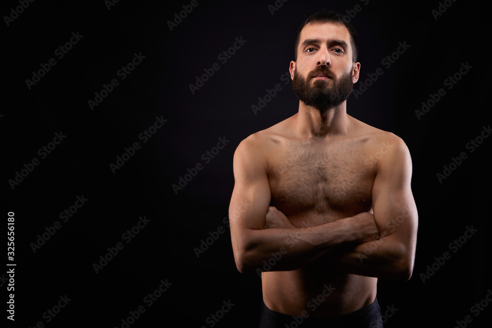 Caucasian young man with a beard, serious, no shirt, muscular body,on black background looking straight ahead with arms crossed, horizontal