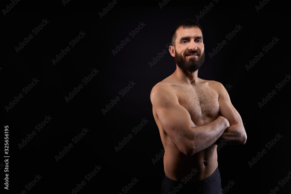 Caucasian young man with a beard, smiling, shirtless, muscular body,on black background looking straight ahead with arms crossed, horizontal