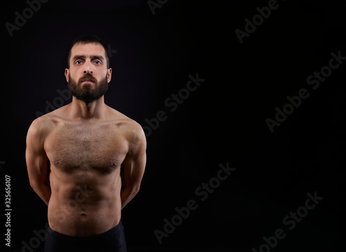 Caucasian handsome young man with a beard, serious, no shirt, muscular body,on black background looking straight ahead, horizontal