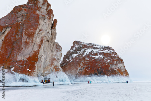 Baikal Lake in the winter. Tourists travel on the ice of frozen lake to the northern edge of Olkhon Island. A group of tourists is photographed against icy rocks of Three Brothers of Cape Sagan-Hushun