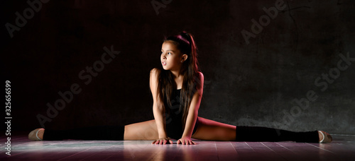 Young smiling girl gymnast in black sport body and special footwear sitting in twine over dark background