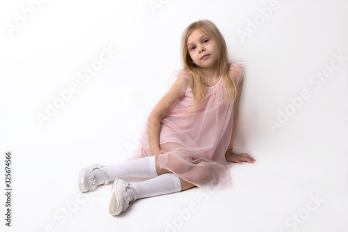 little blonde girl of six seven years in a pink dress and in golf and white shoes is sitting, squatting on a white background