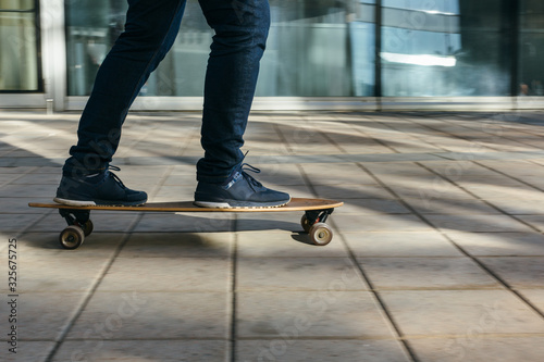 Man in dark blue jeans and sneakers riding on longboard in motion on paving stones or asphalt. Selective focus on skateboard. Sunlight, warm weather. Concept of leisure activity, hobby and sport.
