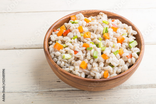 Pearl barley porridge with vegetables in wooden bowl on a white wooden background. Side view, close up.