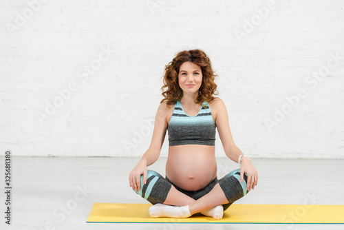 Pregnant girl in sportswear smiling at camera on fitness mat at home