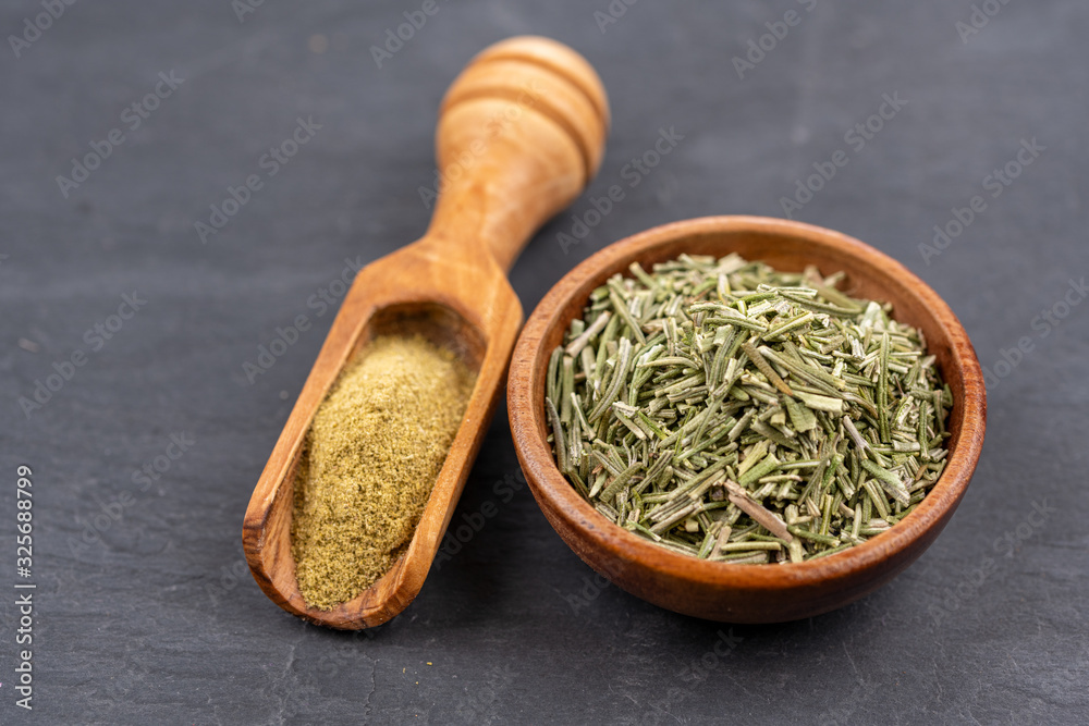A small bowl with whole dried rosemary and a spice shovel with ground rosemary on a black slate background A small bowl with whole dried rosemary and a spice shovel with ground rosemary on a black slate background
