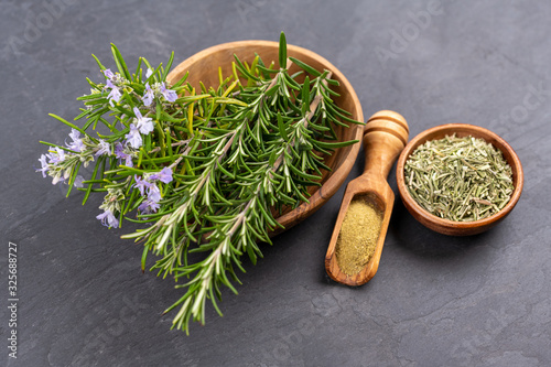 Purple blooming and fresh rosemary twigs in a bowl of olive wood, a small bowl with whole dried rosemary and a spice shovel with ground rosemary on a black slate background