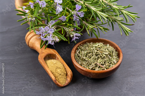 Purple blooming and fresh rosemary twigs in a bowl of olive wood, a small bowl with whole dried rosemary and a spice shovel with ground rosemary on a black slate background
