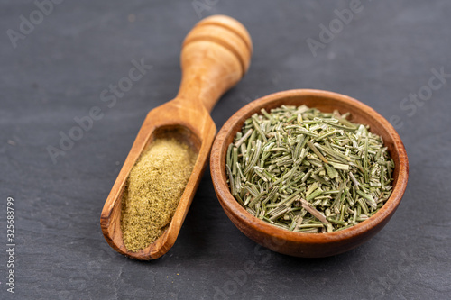 A small bowl with whole dried rosemary and a spice shovel with ground rosemary on a black slate background