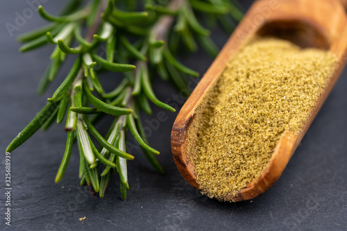 Extreme close-up view of fresh rosemary twigs and a spice shovel with ground rosemary on a black slate background