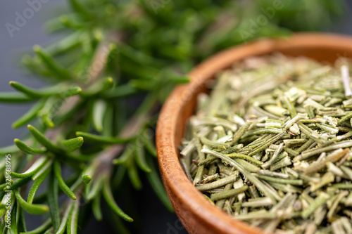 Extreme detail close-up view of fresh rosemary twigs and a wooden bowl with dried rosemary on a black slate background