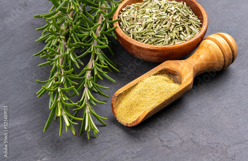 Fresh rosemary twigs, a spice shovel with ground rosemary and a wooden bowl with dried rosemary on a black slate background