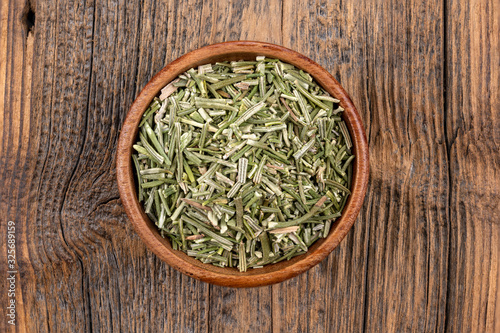 A wooden bowl filled with whole dried rosmary on a rustic wooden background