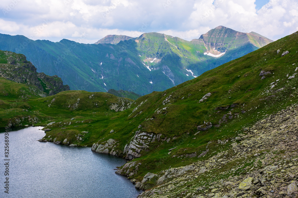 goat lake in the fagaras mountains of romania. popular travel destination. summer nature scenery with green grass and snow. ridge in the far distance beneath a cloudy sky