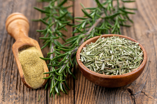 A wooden bowl filled with whole dried rosmary, a spice shovel with ground rosemary and two rosemary twigs on a rustic wooden background