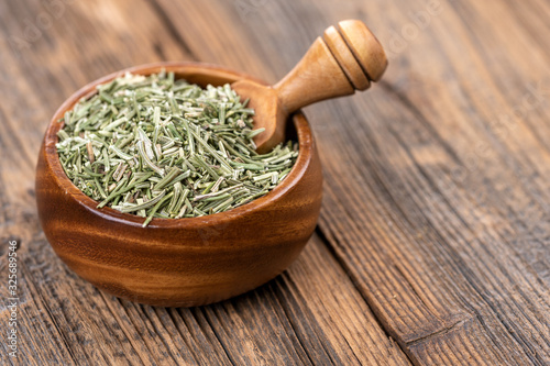 Close-up view of a wooden bowl filled with whole dried rosemary and a small wooden spice shovel on a rustic wooden background