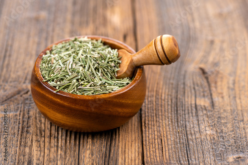 Close-up view of a wooden bowl filled with whole dried rosemary and a small wooden spice shovel on a rustic wooden background