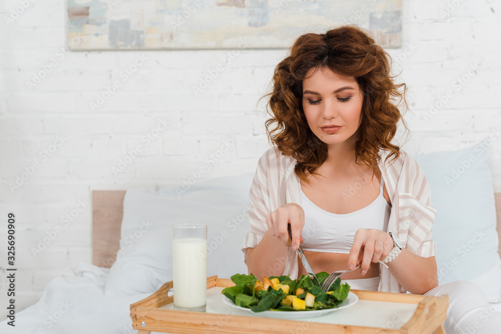 Beautiful woman eating salad near milk on breakfast tray on bed Beautiful woman eating salad near milk on breakfast tray on bed